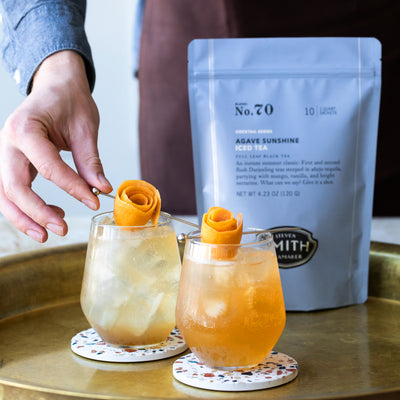 Person placing orange peel garnish on glass filled with Agave Sunshine Spritz with bag of iced tea in background.