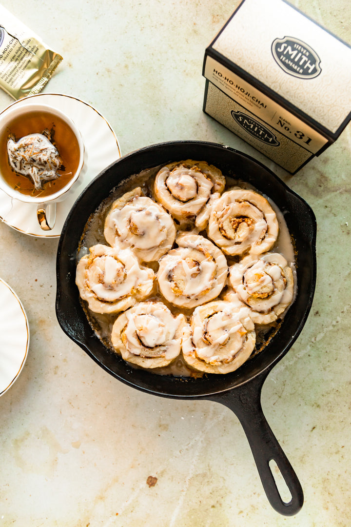 Cast iron pan filled with Ho-Ho Apple Cinnamon Rolls with a brewed cup of tea and box of Ho-Ho-Hoji Chai.