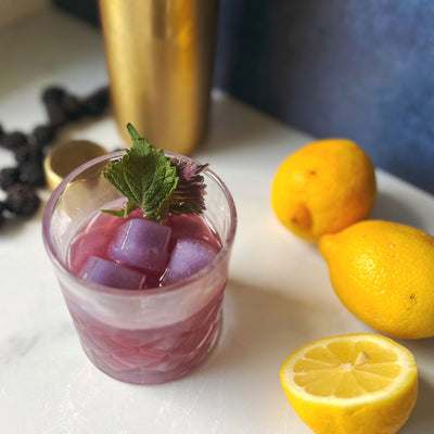 Glass of purple cocktail with shisho leaf garnish with whole and halved lemons on a cutting board with a cocktail shaker in the background.