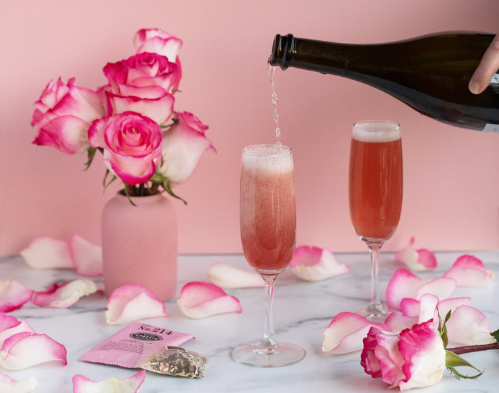 Bottle of prosecco pouring into champagne glass filled with Chivalry cocktail with roses surrounding the glasses.