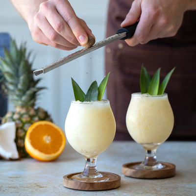 Person grating nutmeg over two glasses of blended Tea Colada with pineapple and orange in the background.
