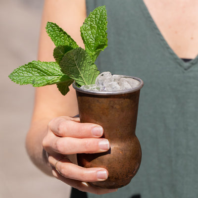 Person holding copper mule cup filled with ice and Mediterranean Mint Julep with large mint sprig garnish.