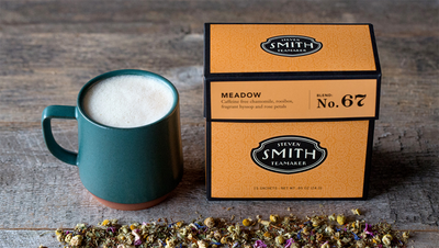 Orange box of Meadow herbal infusion beside a green teacup filled with Meadow Oat Latte.