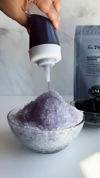 Hand squeezing a squeeze bottle filled with Blackberry Jasmine simple syrup over a glass bowl of shaved ice.