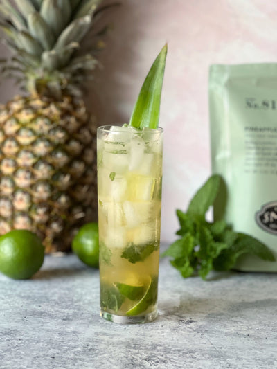 Pineapple Green Mojito in collins glass garnished with pineapple frond with pineapple and limes in background.