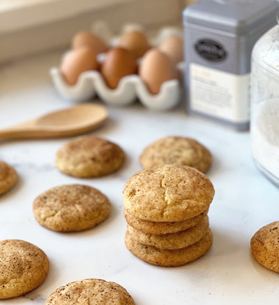 Several Masala Chai Snickerdoodle cookies on a marble countertop with eggs and a tin of tea in the background.