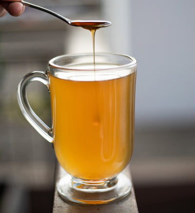 Glass toddy cup filled with Soothe Sayer Toddy and a spoon of honey being drizzled over the top of the beverage.
