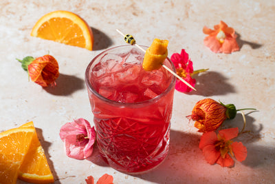 Glass of bright red Summer Fling beverage with orange and floral garnish.