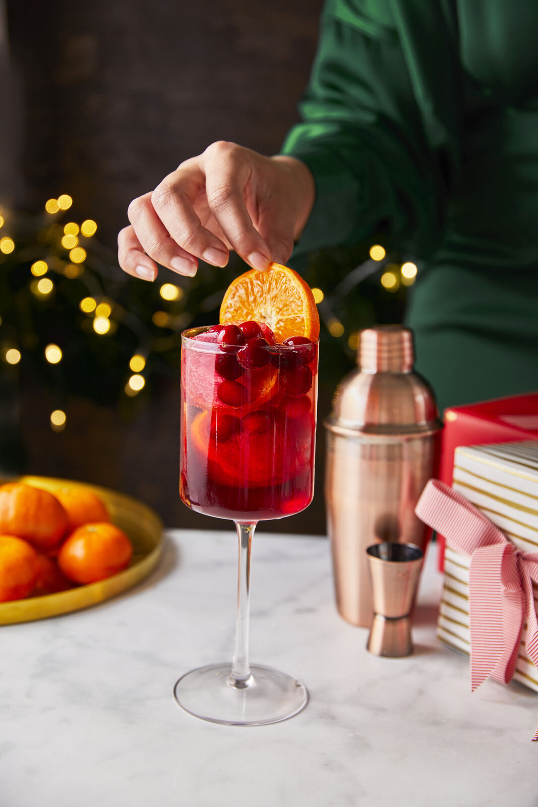 Stemmed wine glass filled with iced Merry Maker's Punch with cranberries and an orange garnish in a holiday scene.