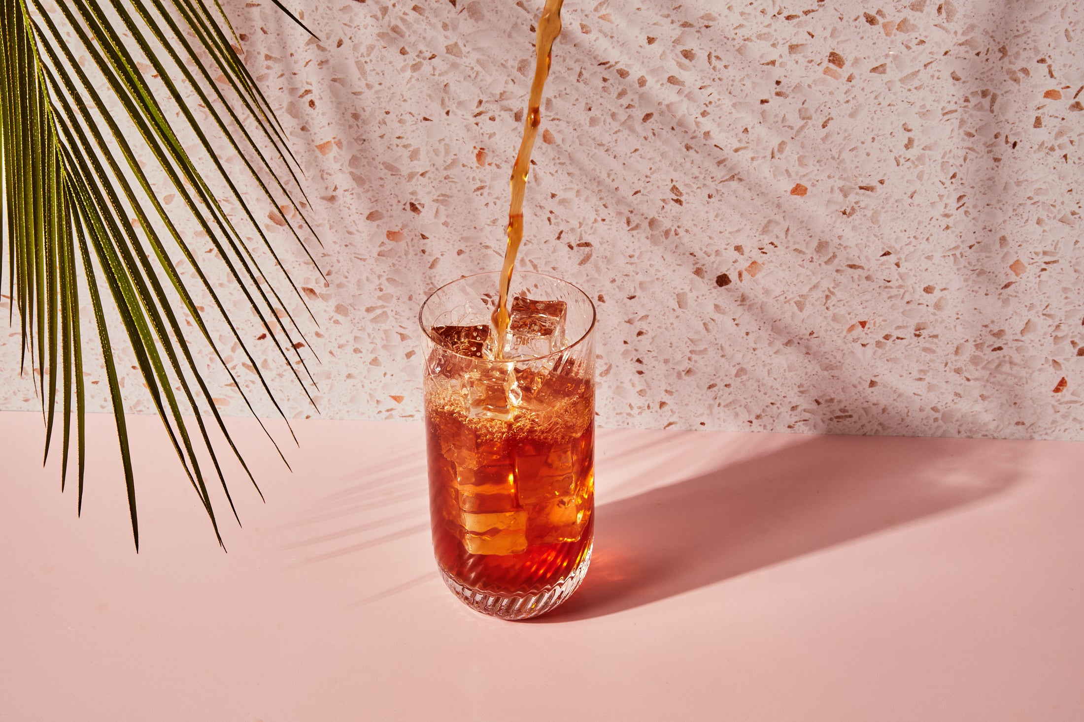 Stream of iced tea being poured into a glass filled with ice with a orange terazzo background.