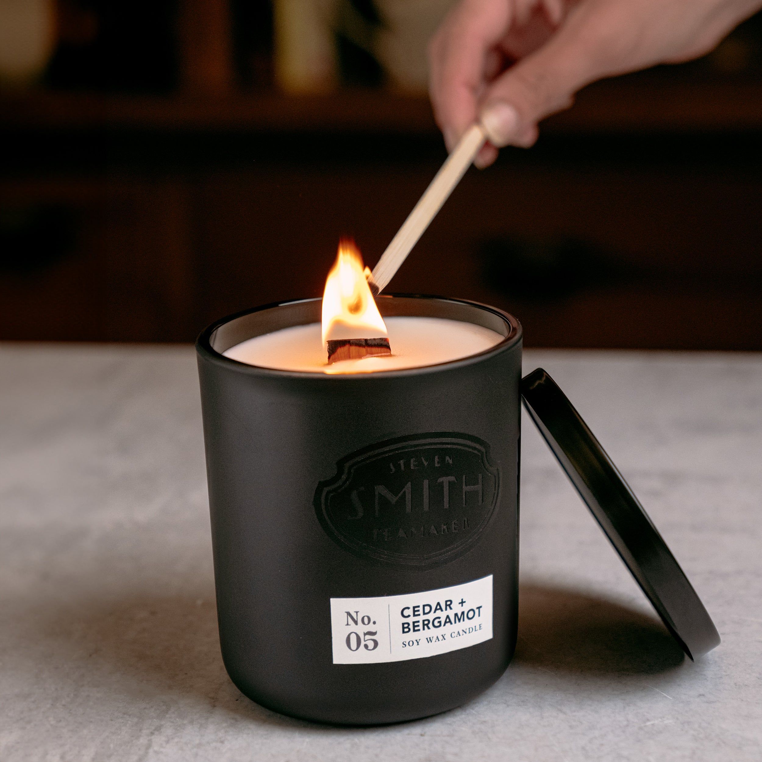 Cedar and bergamot candle with wooden wick being lit by a long wooden match.