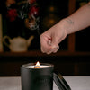 Cedar and bergamot candle with wooden wick being lit by a long wooden match with smoke stream from match.