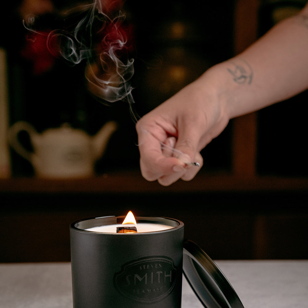 Cedar and bergamot candle with wooden wick being lit by a long wooden match with smoke stream from match.
