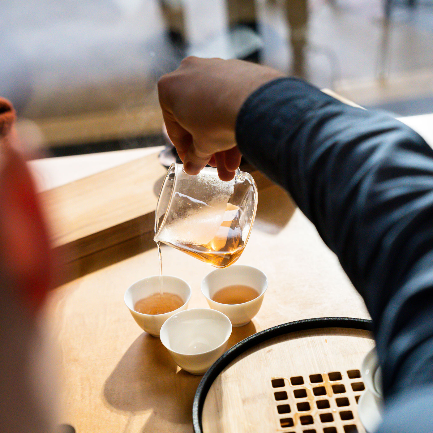 Hand pouring tea into three gaiwan cups beside a gaiwan tray.