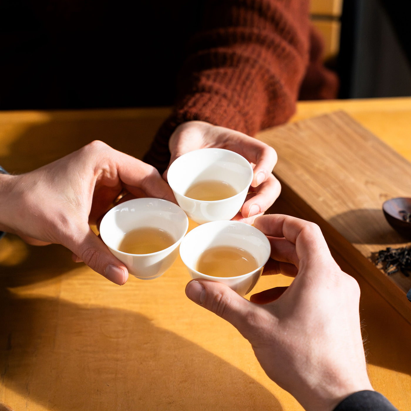 Three hands each holding a small gaiwan cup filled with tea touching to signal a cheers.