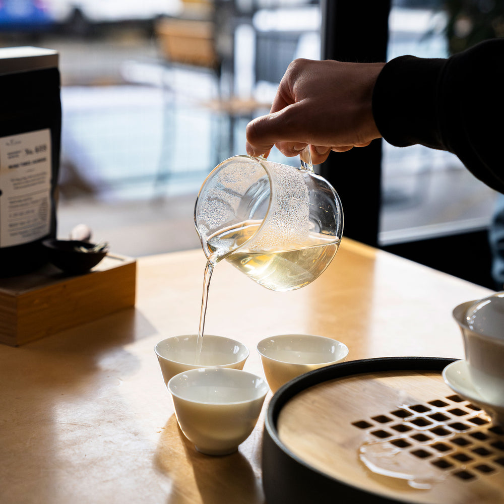Person pouring from a glass teapot into three white ceramic glasses.