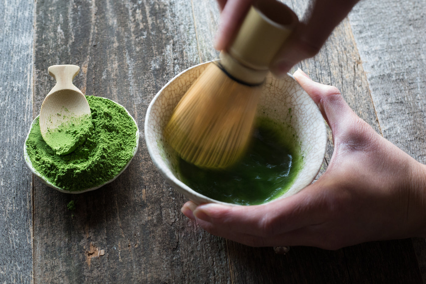 Hand vigorously whisking matcha in a ceramic bowl beside a small pinch pot of matcha powder with a wooden spoon.