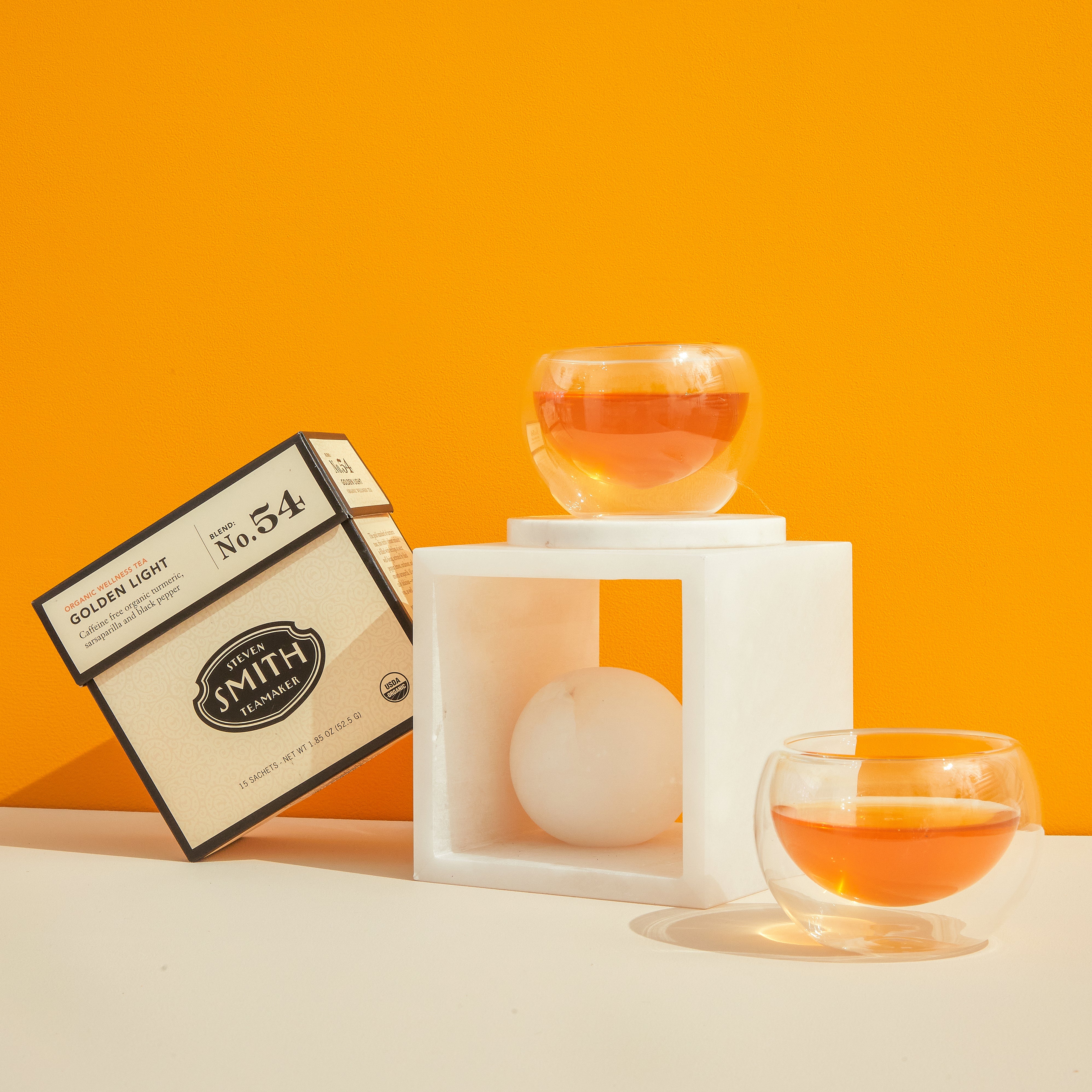 Cream box of Golden Light wellness tea next to two glass cups filled with orange tea.