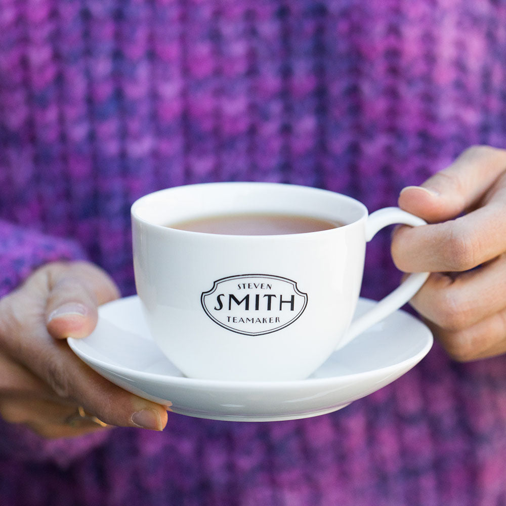 Person in purple sweater holding white porcelain saucer and teacup with black outline of Smith logo shield.