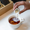 Hand pouring cream into white porcelain cup filled with black tea.