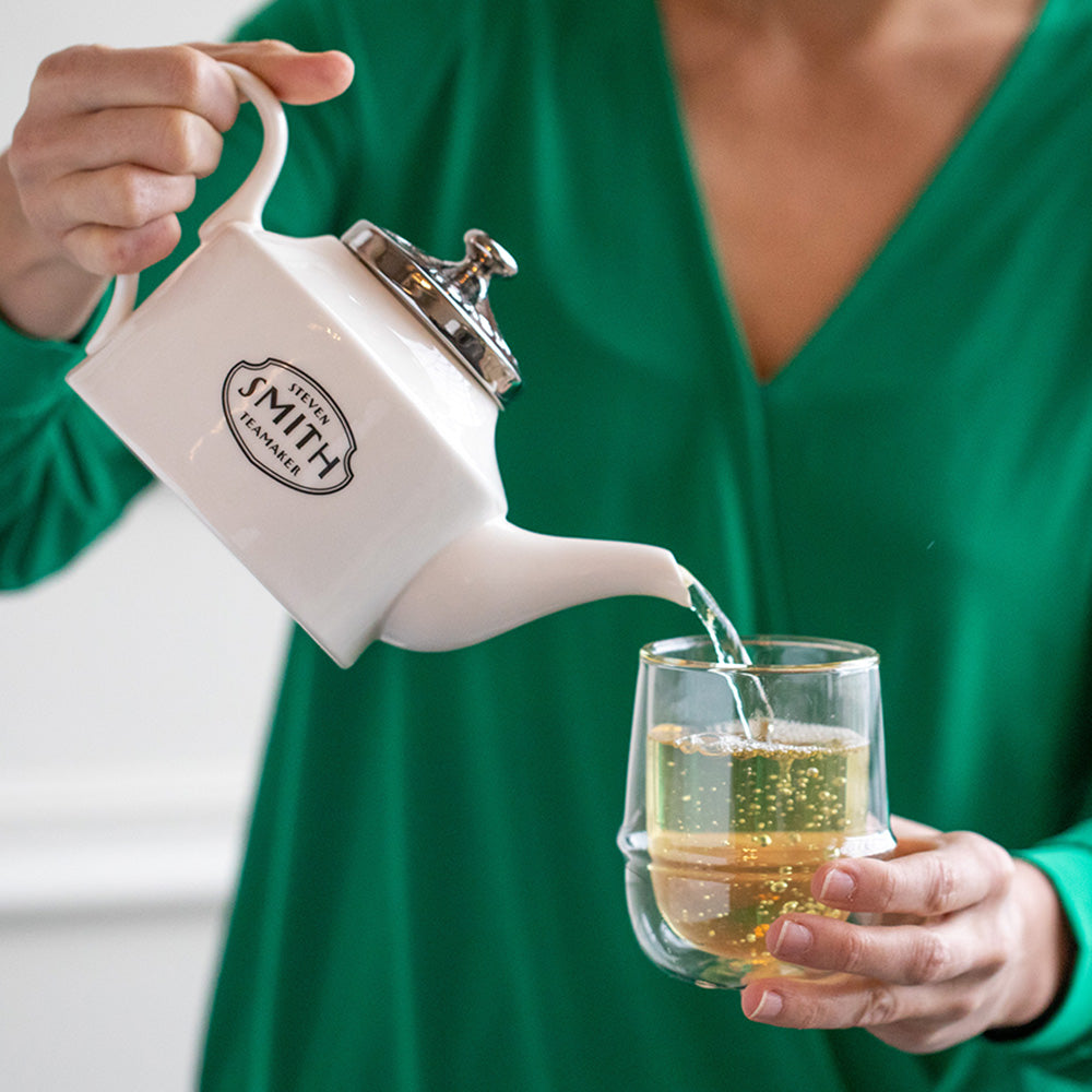 Woman in green shirt pouring from a white teapot into a glass cup.