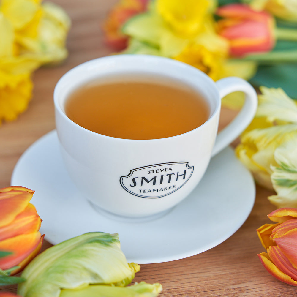 Porcelain teacup and saucer filled with golden liquid and surrounded by orange and yellow flowers.