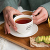 Two hands holding white teacup filled with Portland Breakfast black tea on a tray with sliced bread topped with avocado.