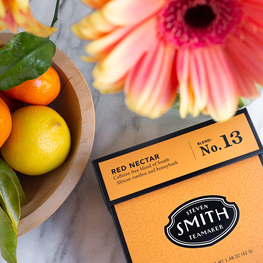 Orange carton of Red Nectar herbal tea alongside bowl of fruit and red flower.