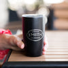 Hand holding black to-go tumbler with silver etched Smith logo shield and clear top.