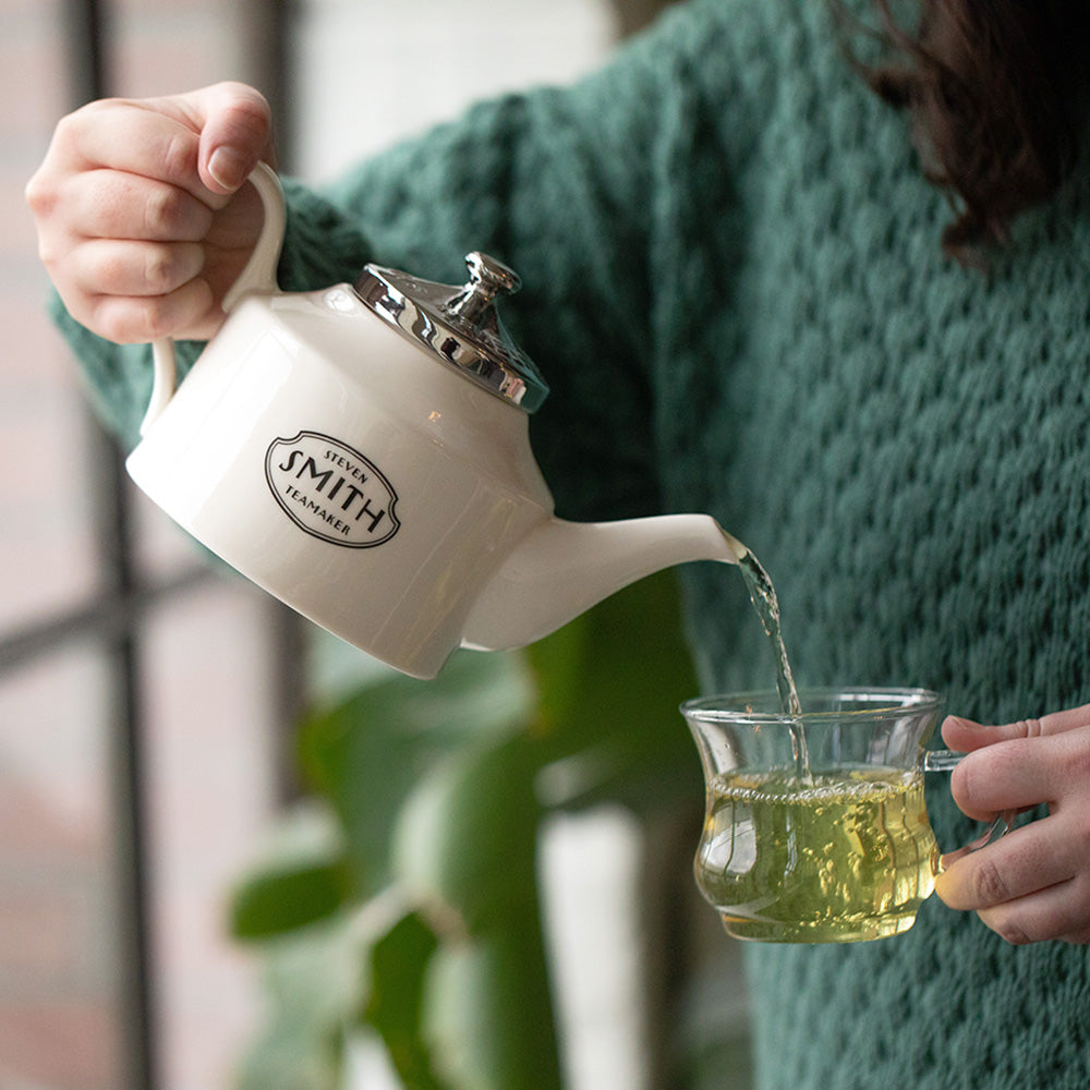 Woman in green sweater pouring White Petal from a white teapot into glass cup.