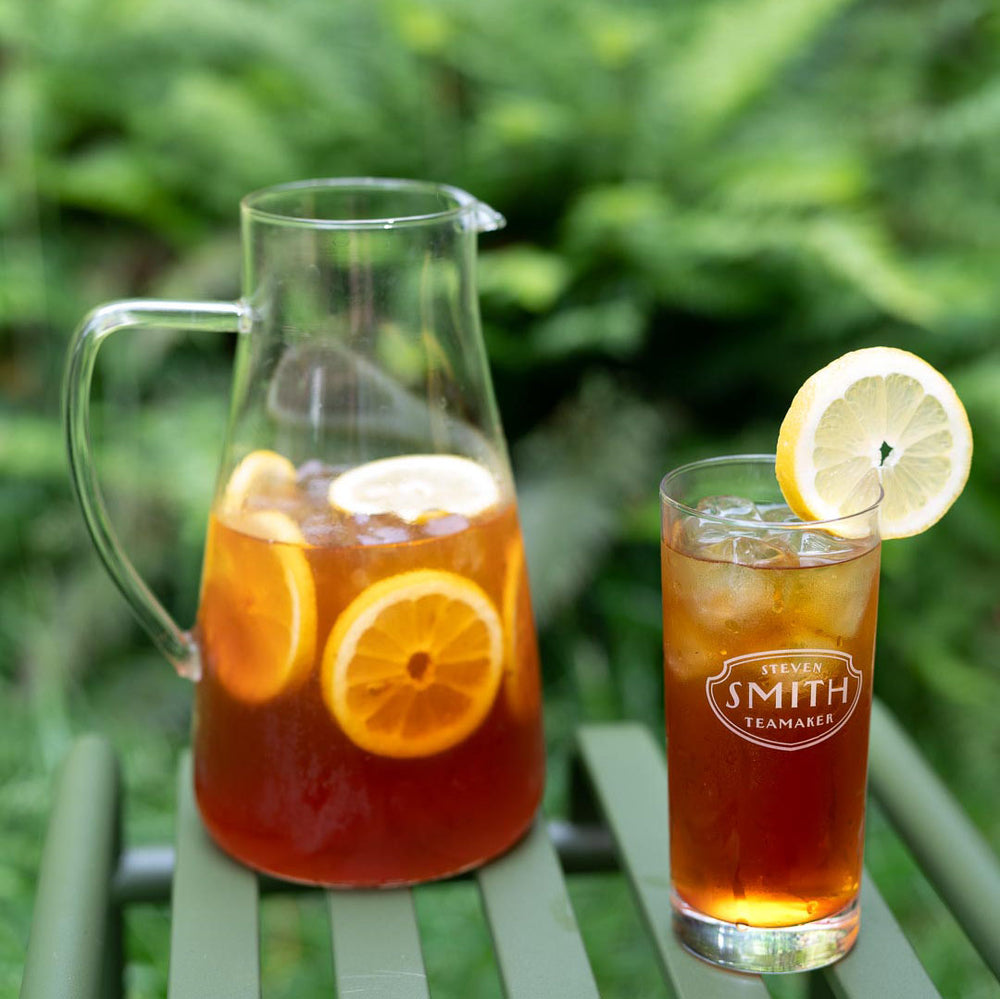 Pitcher filled with lemon wheels and black iced tea next to glass filled with black iced tea on a green table.