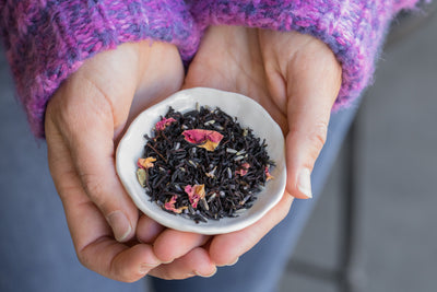 Hands holding small dish filled with loose leaf Black Lavender black tea to show rose petals in blend.