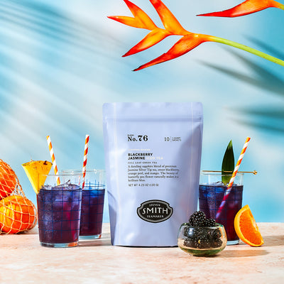 Blackberry Jasmine iced tea pouch with glasses of blue iced tea in front of a tropical background.