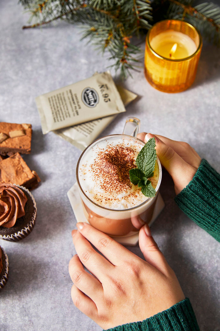 Hands holding glass teacup of Alpine Cocoa with candle and Chocolate Peppermint Pu-erh sachets in background.