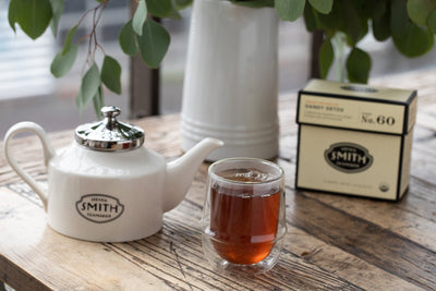Teacup of brewed Dandy Detox wellness tea with Smith teapot and box of Dandy Detox on wooden table.