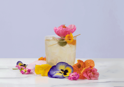 Glass of April Showers mocktail surrounded by flowers with an olive and orange peel garnish with a purple background.