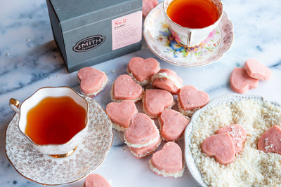 Pile of pink Coconut Kissmet Cookies with two cups of brewed tea and a box of Kissmet Tea.