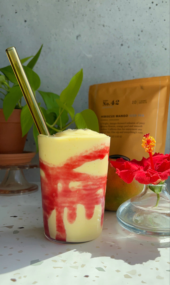 Orange Mango lassi with Hibiscus Mango syrup on inside of glass with straw and pouch of Hibiscus Mango iced tea.
