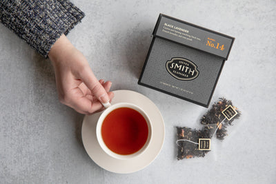 Hand reaching for a teacup filled with Black Lavender tea and a box of Black Lavender tea beside two unwrapped sachets of tea.