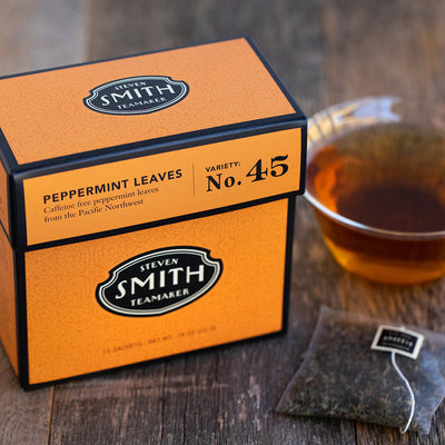 Orange box of Peppermint Leaves herbal infusion, an unwrapped sachet and a glass teacup of brewed tea on wooden surface.