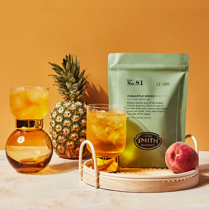 Pouch of Pineapple Green iced tea on a whicker tray with peach, pineapple and glasses of iced tea.