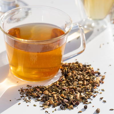 Glass teacup of brewed Soothe Sayer wellness blend beside pile of loose leaf Soothe Sayer.