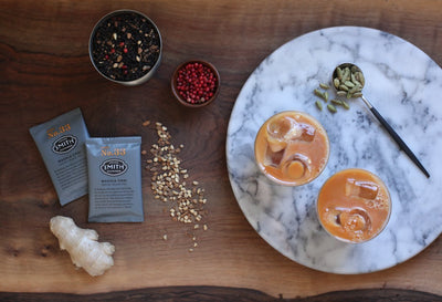 Two iced chai lattes on a marble board with sachets of Masala Chai, ginger root, peppercorns and loose tea beside.