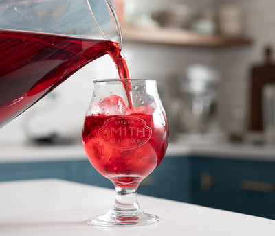Pitcher pouring red hibiscus mango iced tea into a snifter glass filled with ice.