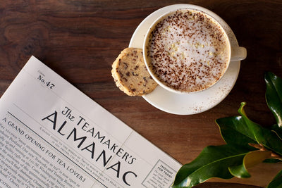 Black Tea Latte with cocoa powder garnish on a saucer with a cookie beside a newspaper.