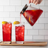 Person pouring red tea from glass pitcher into two glasses filled with ice.