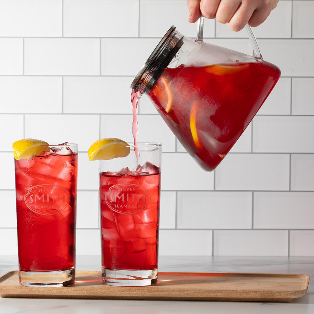 Person pouring red tea from glass pitcher into two glasses filled with ice.