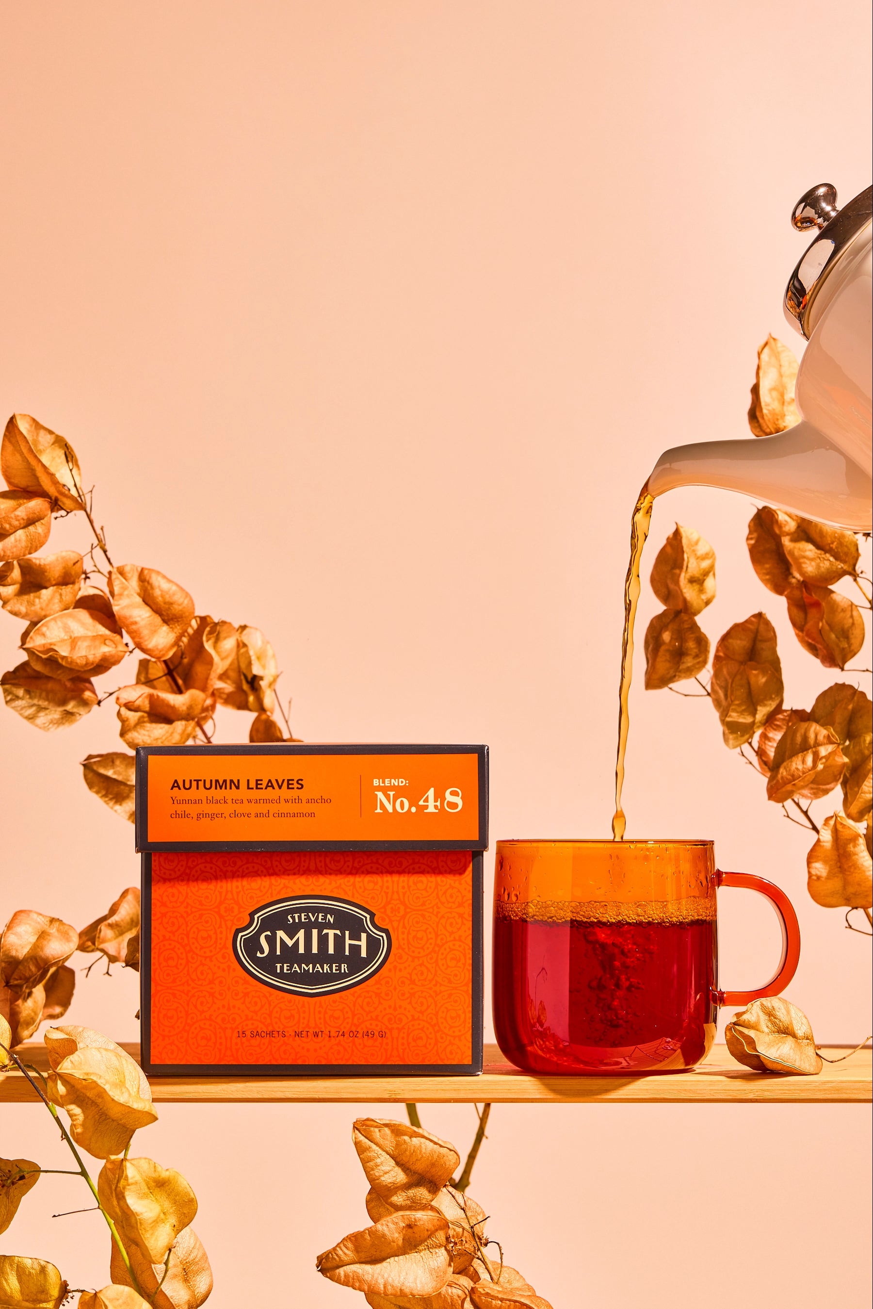 A box of  Smith Tea Autumn Leaves No.45 tea sits on a wooden shelf next to a glass mug filled with brewed tea, with dried leaves and a teapot in a warm peach studio setting.
