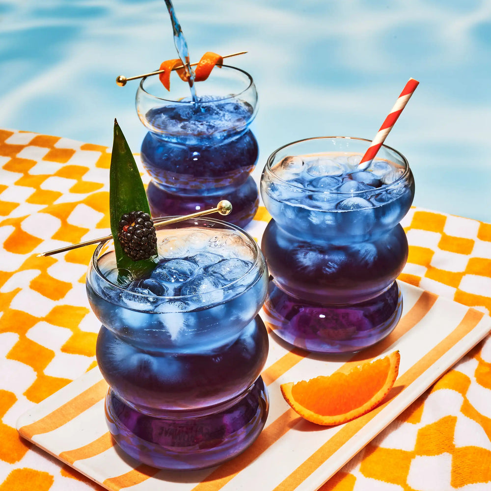 Three glasses of Blackberry Jasmine iced tea in wavy glasses with festive garnishes on an orange and white checkered pool towel.