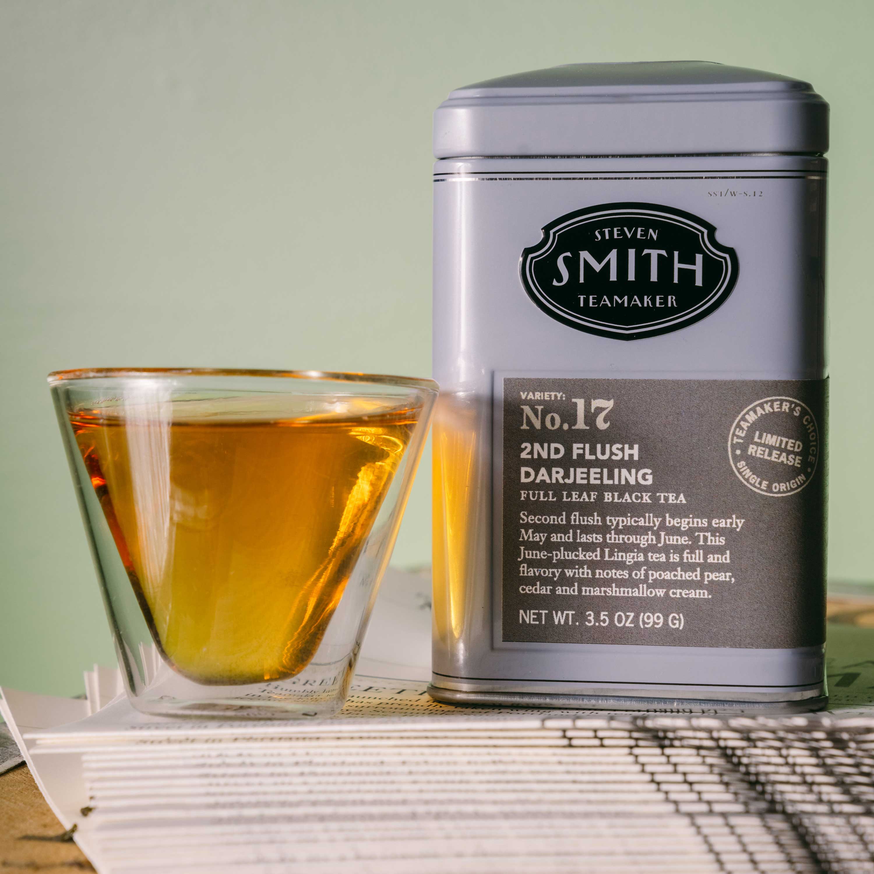 A silver tin of Steven Smith Teamaker Darjeeling No. 17 full leaf black tea stands next to a clear glass cup filled with amber tea, set on a stack of papers against a soft sage green background.